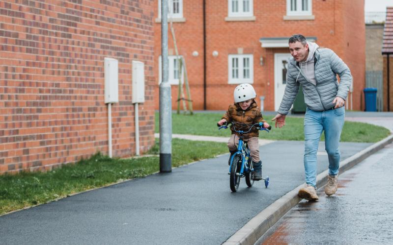 A father walks alongside his young son as he rides his bike through a housing estate.