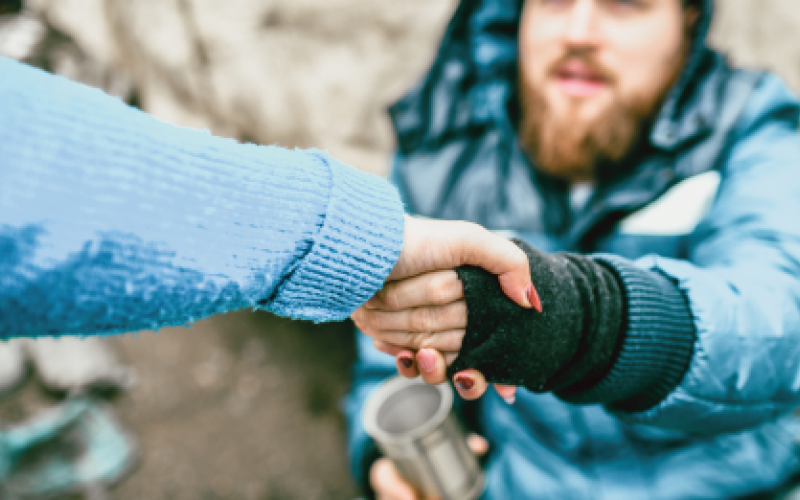 Photograph of an outstretched arm grasping the arm of a seated, bearded homeless man