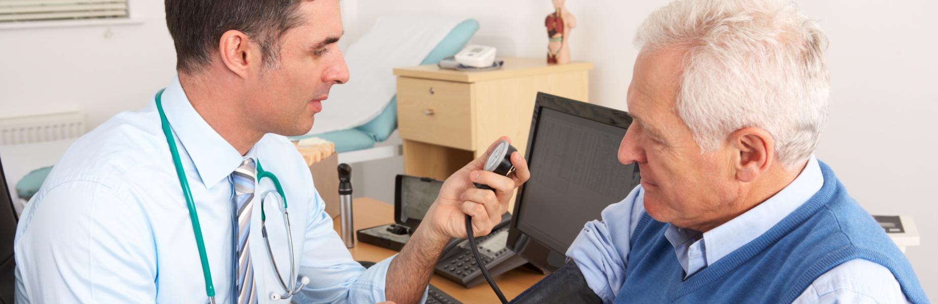 A healthcare professional with a stethoscope checks a patient’s blood pressure using a monitor in a medical office setting. A computer screen and medical equipment are visible on the desk